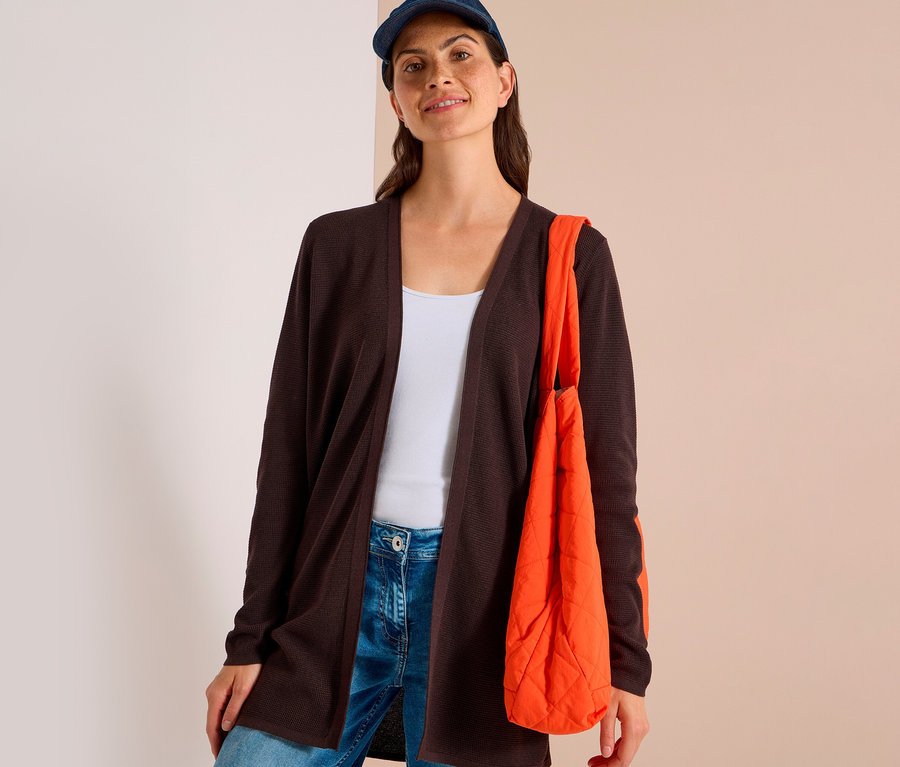 Frau trägt eine dunkelbraune CECIL Strickjacke über einem weißen Tanktop und blauen Jeans, mit blauer Kappe und orangefarbener Stepptasche.