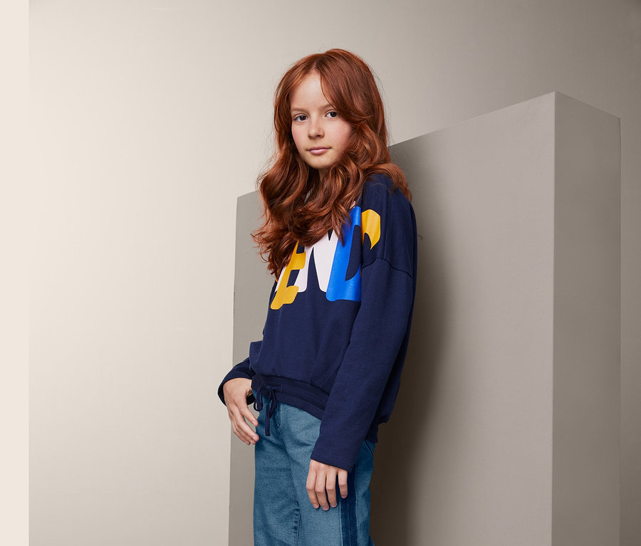Ein junges Mädchen mit gewellten, roten Haaren posiert in einem dunkelblauen Sweatshirt, einer Jeans-Culotte mit Streifen und weißen Turnschuhen.
