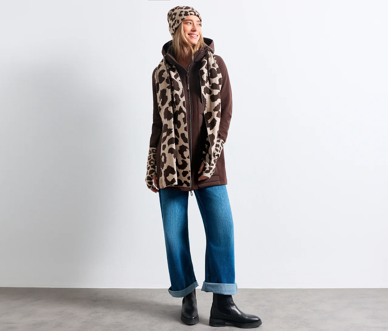 Eine lächelnde Frau trägt eine braune Kapuzenjacke, blaue Jeans, schwarze Stiefel und eine Street One Strickmütze mit Leopardenmuster, passend zum Schal und den Ärmeln.