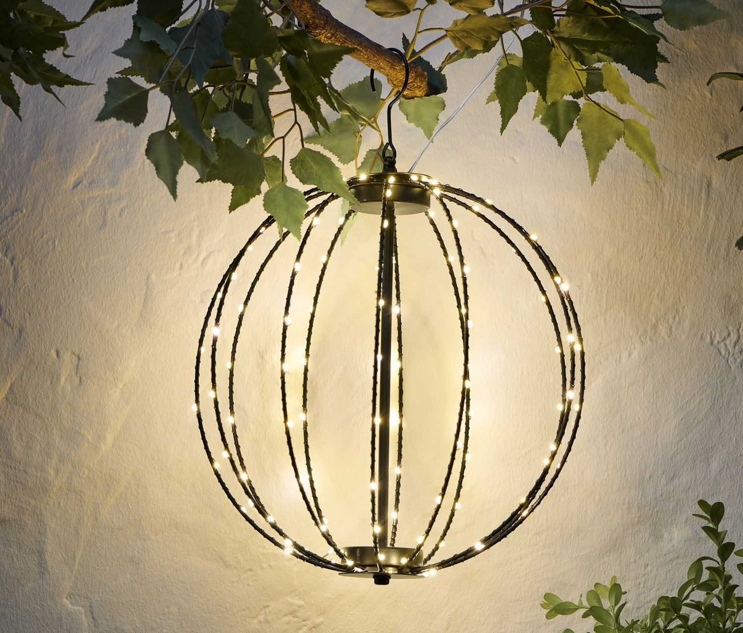 Eine LED-Kugel aus Metall hängt an einem Ast vor einer weißen Wand.