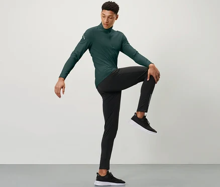 Mann in grünem Sportoberteil und schwarzen Windprotection-Lauftight hält ein Knie mit einer Hand vor der Brust.