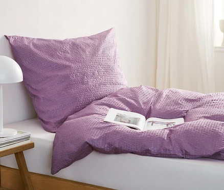 Lila Seersucker-Bettwäsche liegt auf einem Bett mit einem offenen Buch. Auf dem Nachttisch stehen eine Lampe und Bücher.