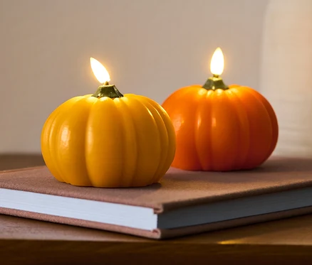 Zwei LED-Echtwachskerzen »Kürbis« stehen auf einem Buch auf einem Holztisch.