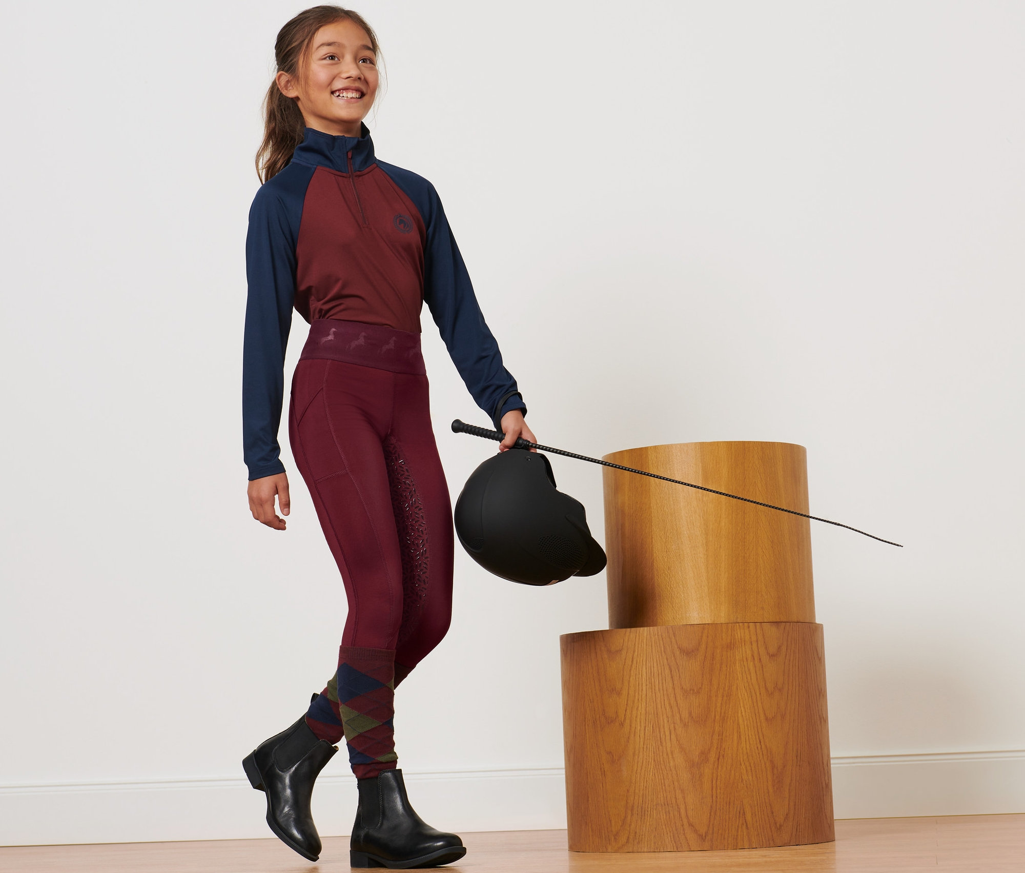 Mädchen hält Reithelm und Peitsche, trägt eine rote Kinder-Grip-Vollbesatz-Reittight.