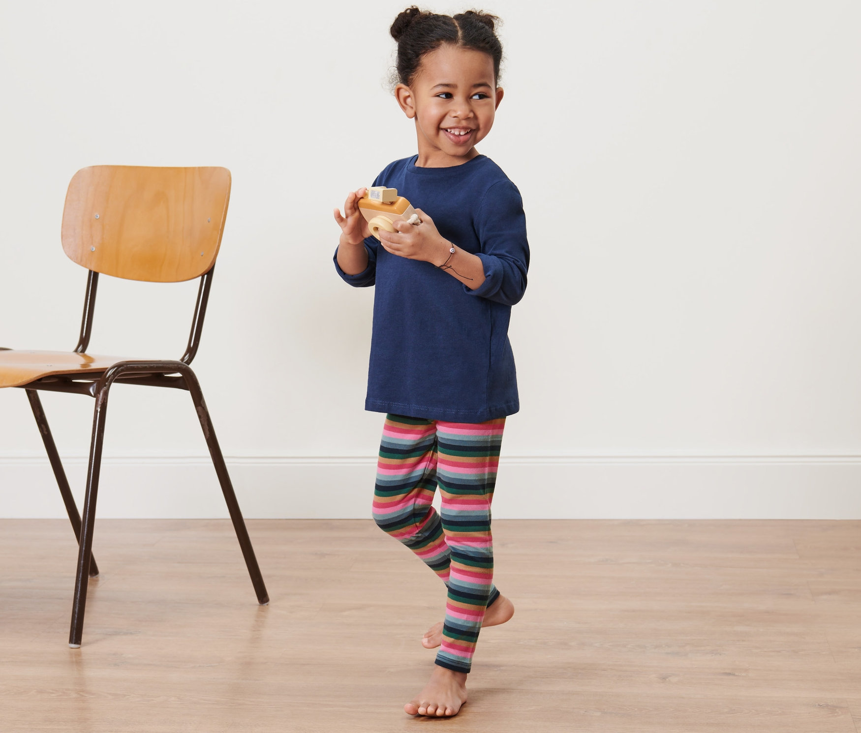 Ein lächelndes kleines Mädchen hält ein Holzspielzeug in ihren Händen und trägt angeraute Kleinkind-Leggings.