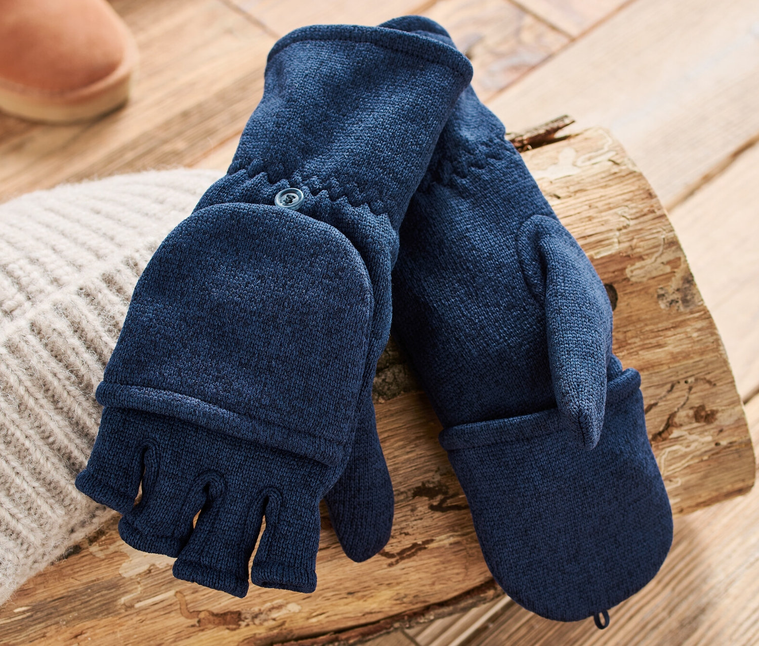 Dunkelblaue Strickfleece-Klappfäustlinge liegen auf einem Holzscheit neben einer Strickmütze und braunen Stiefeln.