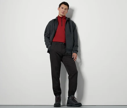 Ein Mann steht und trägt eine Thermohose mit Gürtel, eine Zip-Jacke und Wanderschuhe.