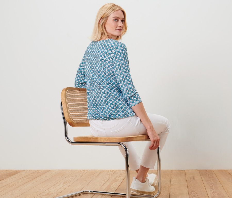 Frau sitzt auf einem Stuhl und trägt ein Blusenshirt mit Alloverprint.