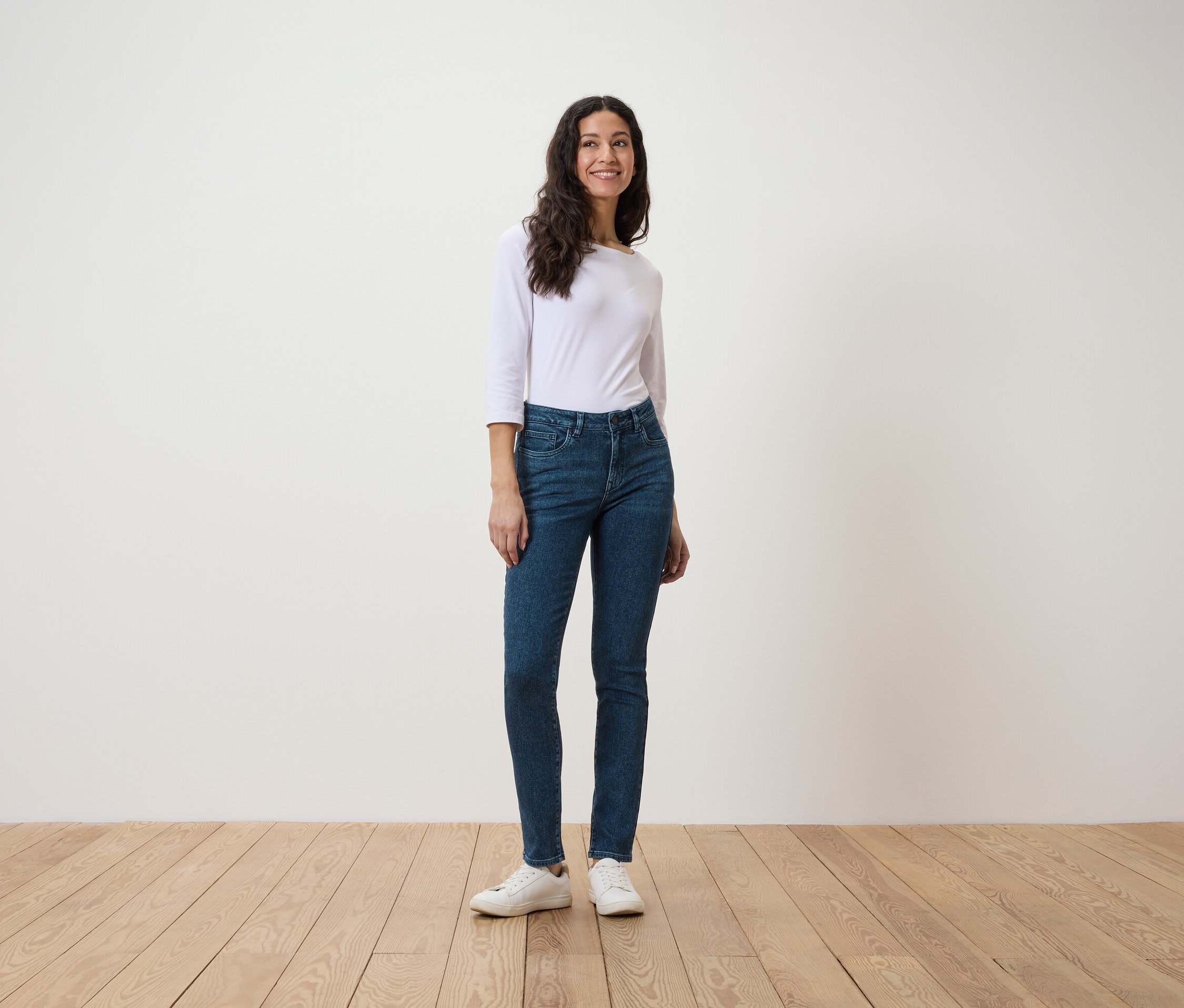 Eine Frau steht auf einem Holzboden und trägt ein weißes Dreiviertelarmshirt und blaue Jeans.