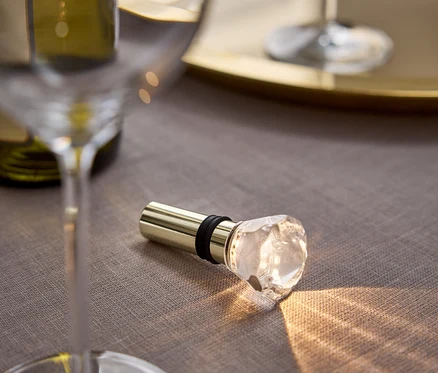 Auf einem grauen Tuch liegen ein goldener Weinstopfen mit Kristallkopf, daneben ein Glas und eine Flasche Wein.