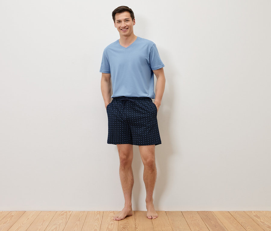 Ein Mann steht barfuß mit den Händen in den Taschen seiner marineblauen, gepunkteten Shorts da und trägt ein hellblaues T-Shirt vor einer weißen Wand.