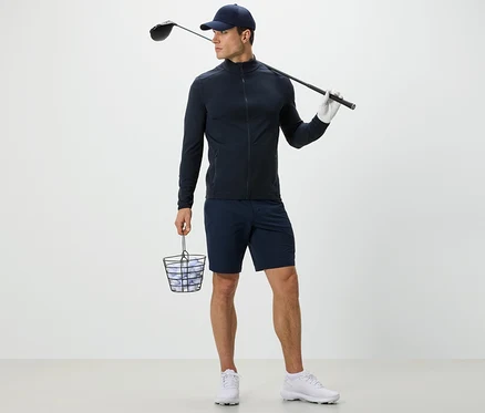Mann in marineblauer Funktionsjacke hält Korb mit Golfbällen und Golfschläger.