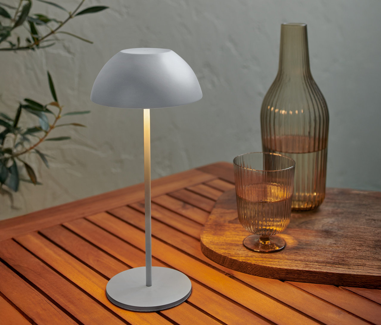 Eine graue Lampe steht auf einem Holztisch neben einer Flasche und einem Glas Wasser.