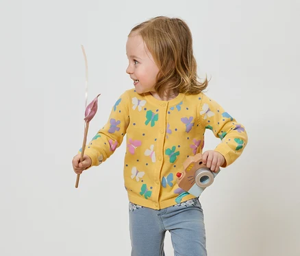 Mädchen mit gelber Schmetterlingsjacke hält eine Spielzeugkamera und eine Blume.