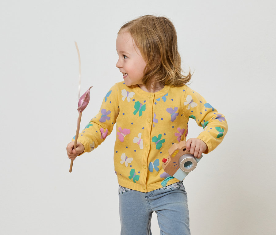 Mädchen mit gelber Schmetterlingsjacke hält eine Spielzeugkamera und eine Blume.