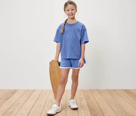 Mädchen hält ein Skateboard, trägt ein blaues T-Shirt und blaue Shorts.