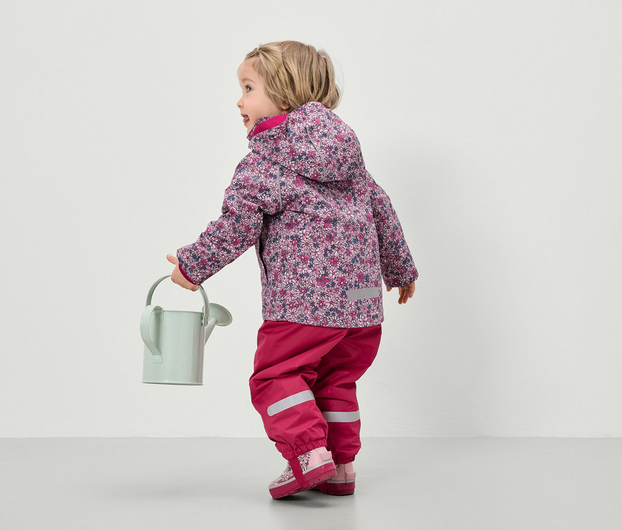 Ein Kind in einer geblümten Jacke und einer pinken Hose hält eine Gießkanne.