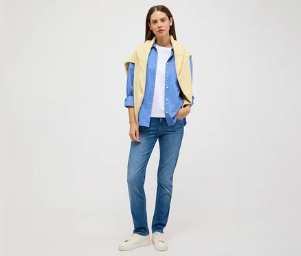 Frau steht vor hellem Hintergrund und trägt Angels Jeans »Cici Push-Up«, Slim Fit, ein blaues Hemd, ein weißes T-Shirt und einen gelben Pullover.
