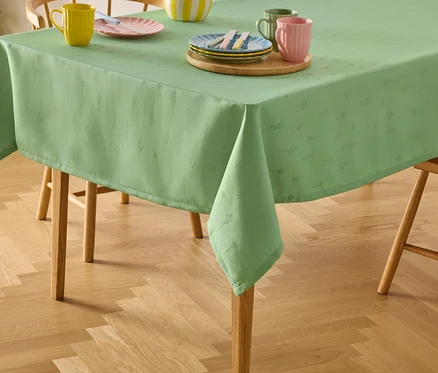 Grüne Tischdecke mit Blumenmuster auf einem Holztisch mit Stühlen, darauf Teller und Tassen.
