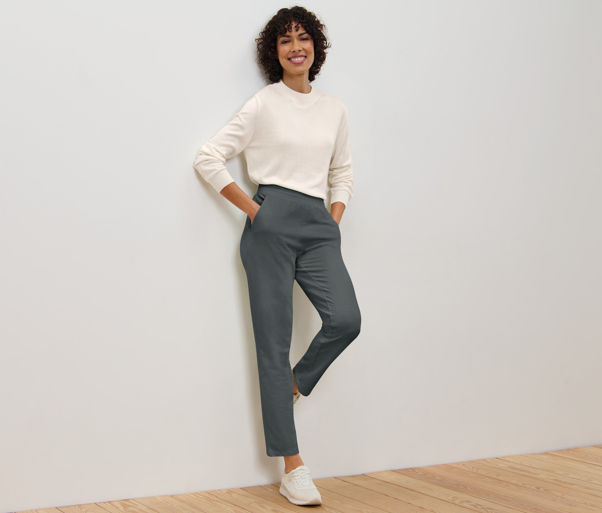 Eine Frau posiert an einer Wand, sie trägt eine graue Joggpants und einen Pullover.