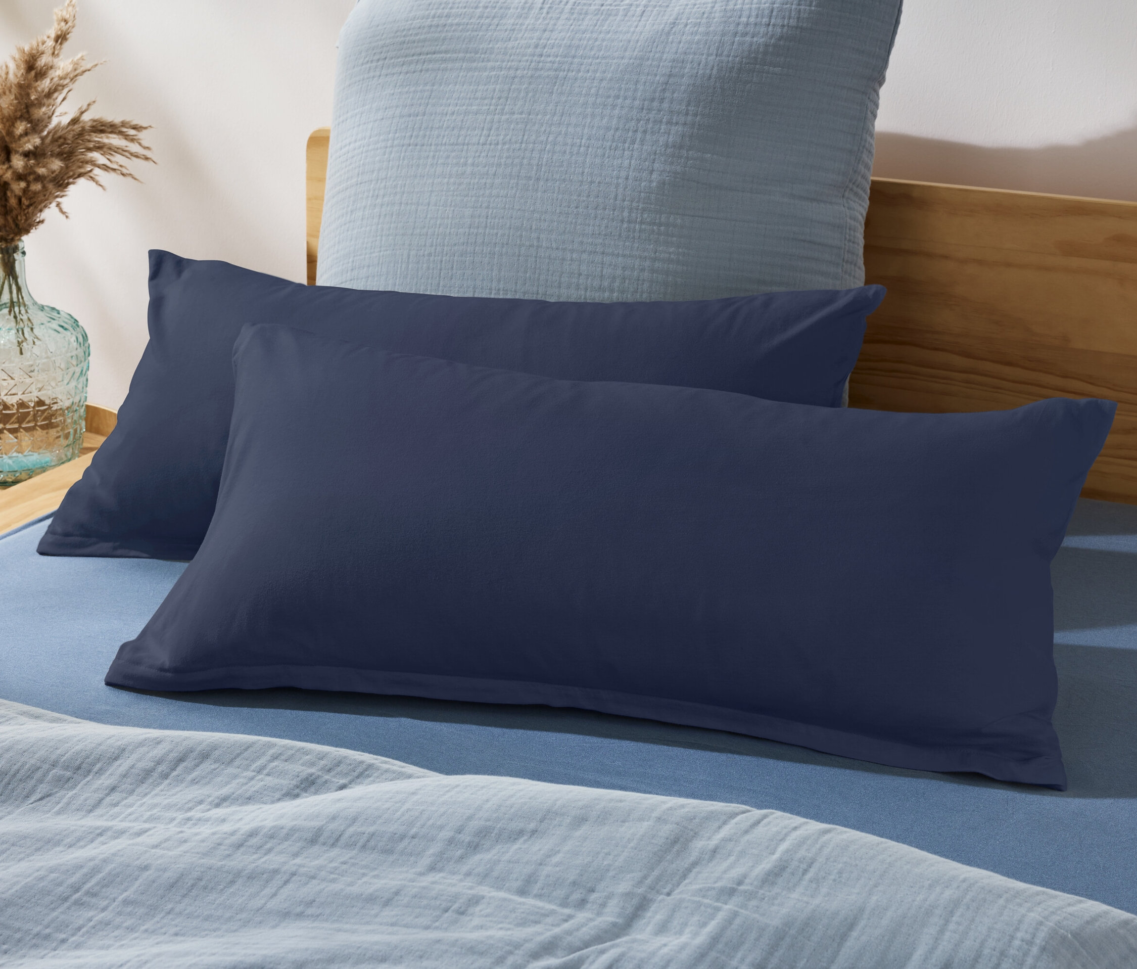 Zwei marineblaue Kissen auf einem blauen Bett mit hellblauer Decke und Vase mit Pampasgras.