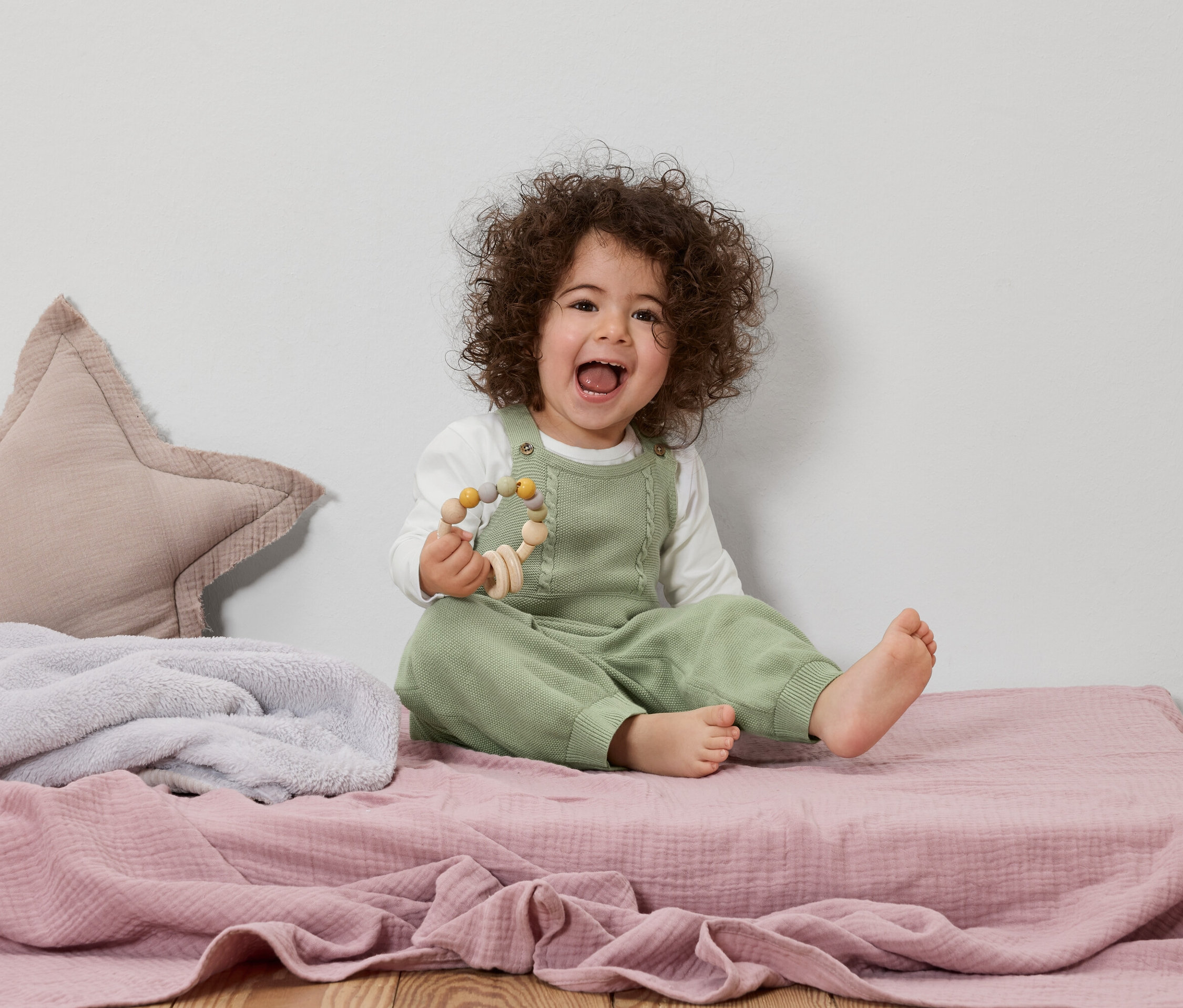 Ein lachendes Kleinkind mit lockigem Haar sitzt auf einer rosa Krabbeldecke und hält ein Holzspielzeug.