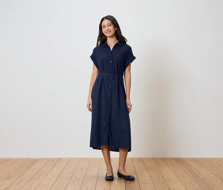 Eine Frau steht vor einer Wand und trägt ein dunkelblaues Hemdkleid mit Gürtel und Ballerinas.