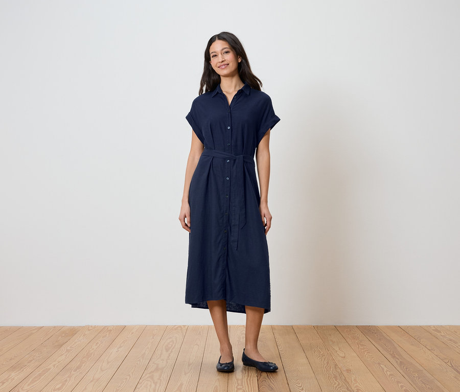 Eine Frau steht vor einer Wand und trägt ein dunkelblaues Hemdkleid mit Gürtel und Ballerinas.