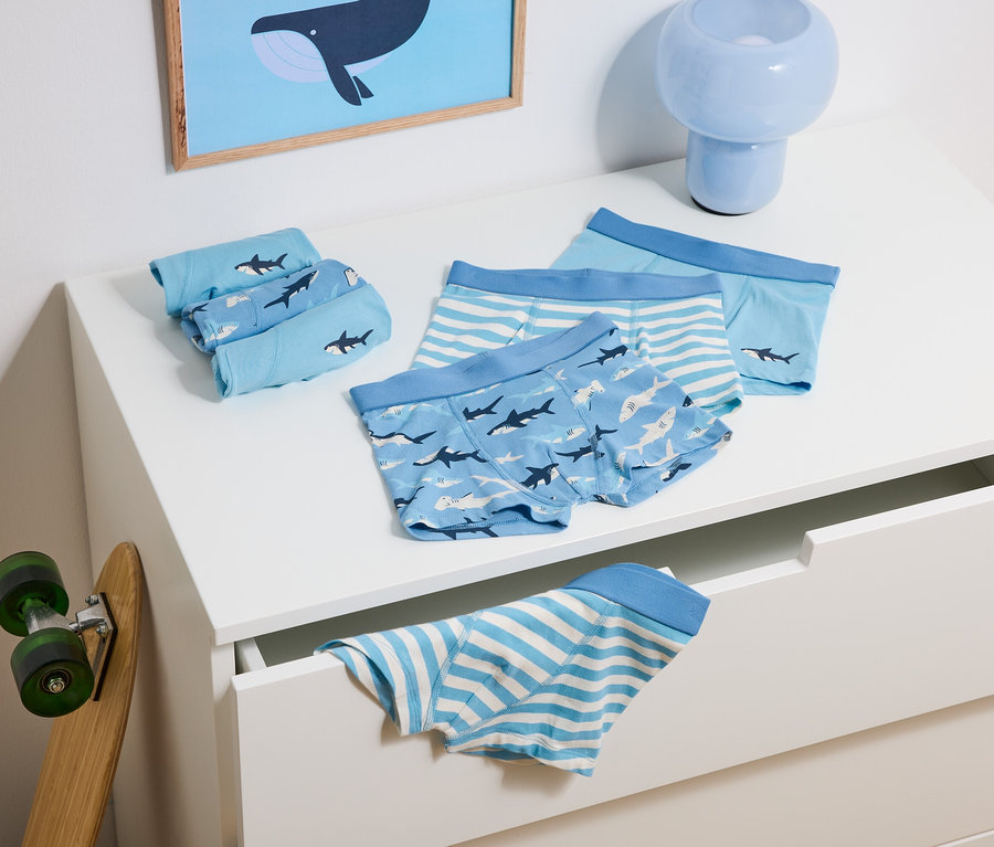 Auf einer Kommode liegen blaue Kinderunterhosen mit Hai- und Streifenmuster, daneben ein Skateboard und eine blaue Lampe.