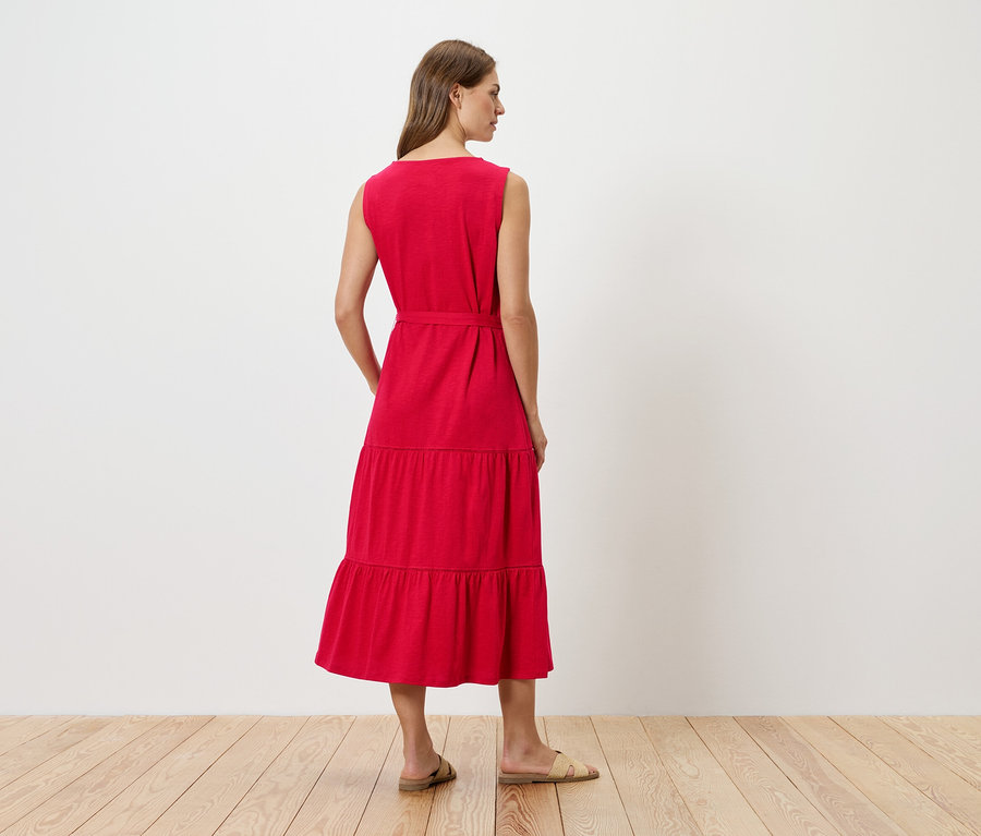 Frau von hinten in einem roten, ärmellosen, gestuften Midikleid mit Gürtel, dazu flache Sandalen.