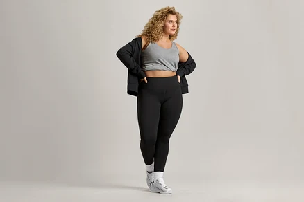 Frau mit lockigem Haar trägt ein graues Tanktop, eine schwarze Jacke und schwarze Leggings.