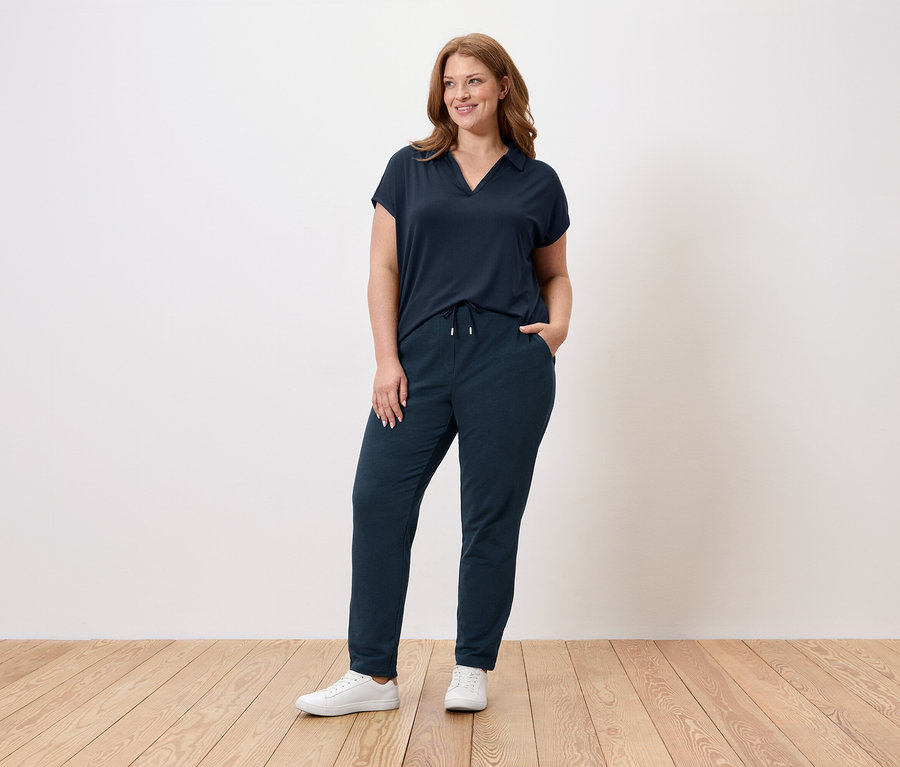 Eine Frau steht vor einer Wand, sie trägt ein dunkelblaues T-Shirt, eine dunkelblaue Hose und weiße Sneaker.
