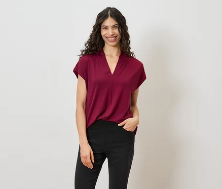 Frau mit lockigem, dunklem Haar trägt ein weinrotes T-Shirt und schwarze Hosen vor einem weißen Hintergrund.