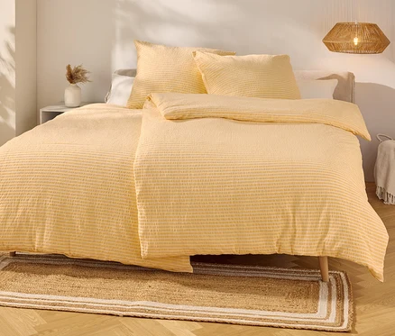 Gelb gestreifte Bettwäsche liegt auf einem Bett mit zwei Kissen. Ein Teppich liegt davor.