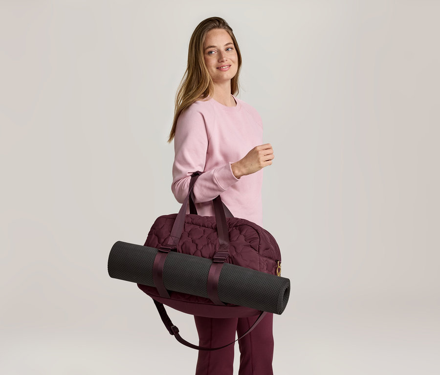 Frau mit rosa Pullover und weinroter Hose, die eine weinrote Sporttasche mit einer schwarzen Yogamatte über der Schulter trägt.