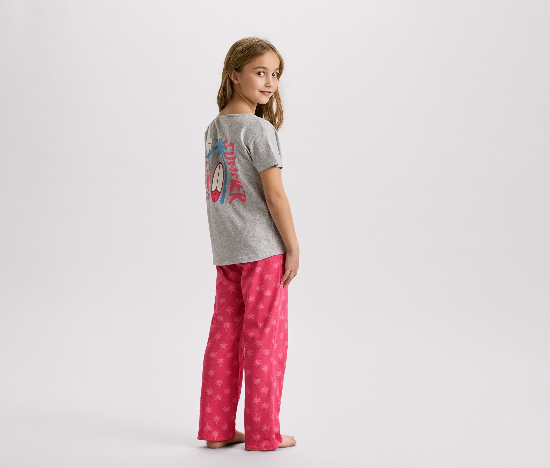 Ein junges Mädchen steht in einem grauen T-Shirt mit einem Sommerdesign und einer rosa Hose mit Palmen.