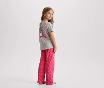 Ein junges Mädchen steht in einem grauen T-Shirt mit einem Sommerdesign und einer rosa Hose mit Palmen.