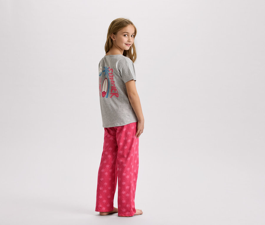 Ein junges Mädchen steht in einem grauen T-Shirt mit einem Sommerdesign und einer rosa Hose mit Palmen.