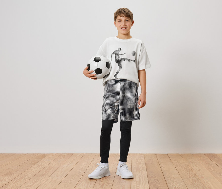 Ein Junge hält einen Fußball in seiner linken Hand. Er trägt ein T-Shirt mit einem Fußballspieler, Shorts und Leggings und weiße Turnschuhe.