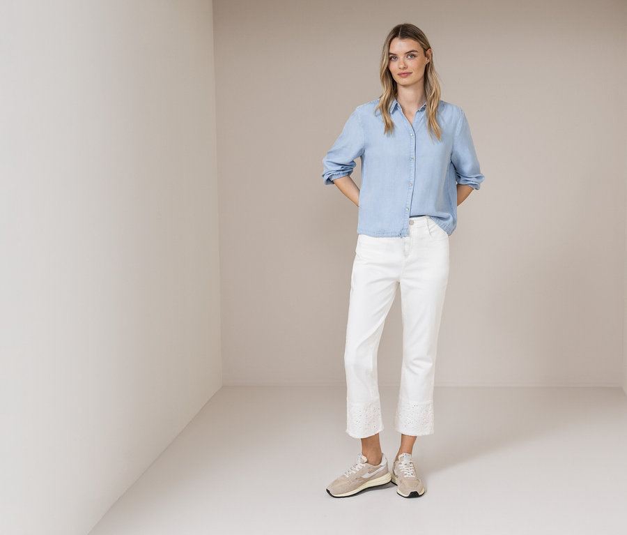 Eine Frau steht in einem Raum und trägt ein hellblaues Hemd, weiße Jeans mit Spitze und beigefarbene Turnschuhe.