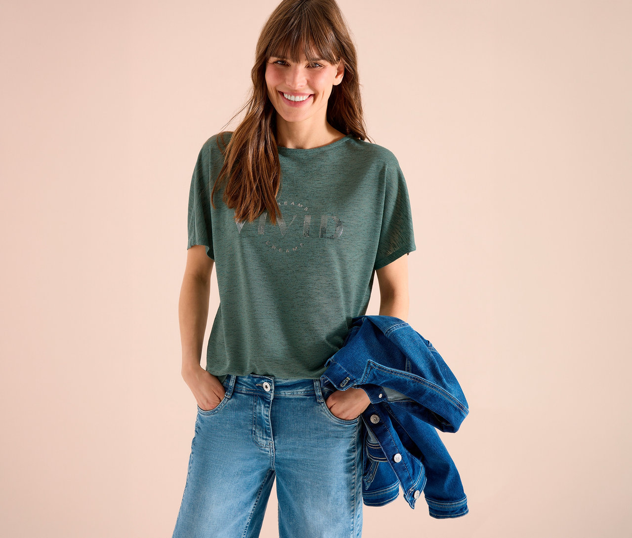 Lächelnde Frau mit braunen Haaren, grünem T-Shirt mit Aufdruck, blauen Jeans und Jeansjacke über dem Arm vor beigem Hintergrund.