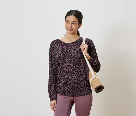 Frau mit einem lila-schwarzen Leopardenmuster-Oberteil und lila Leggings, die eine zusammengerollte Yogamatte über der Schulter trägt.