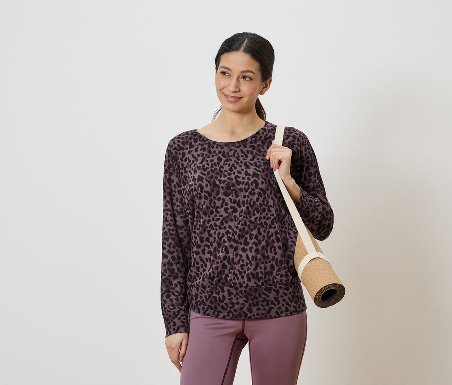 Frau mit einem lila-schwarzen Leopardenmuster-Oberteil und lila Leggings, die eine zusammengerollte Yogamatte über der Schulter trägt.