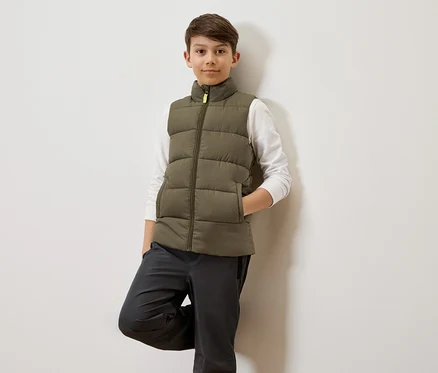 Ein Junge mit braunen Haaren lehnt an einer weißen Wand und trägt eine olivgrüne Steppweste über einem weißen Langarmshirt und graue Hosen.