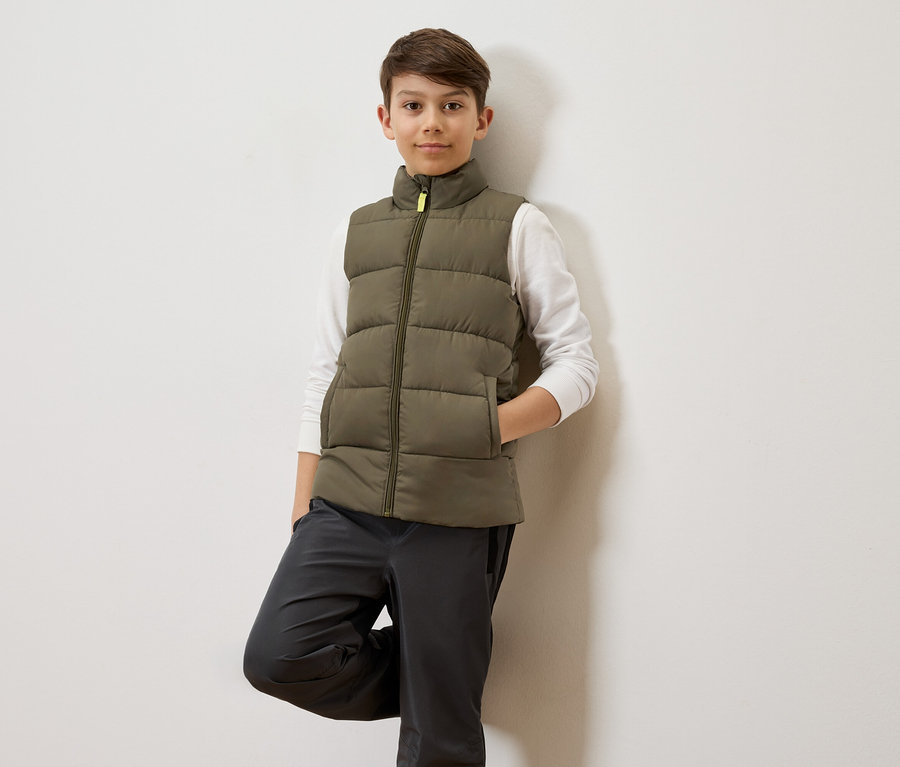 Ein Junge mit braunen Haaren lehnt an einer weißen Wand und trägt eine olivgrüne Steppweste über einem weißen Langarmshirt und graue Hosen.