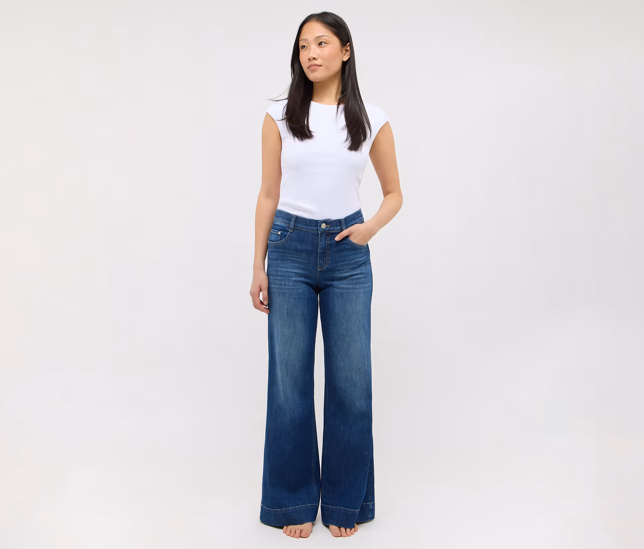Eine Frau steht in einem weißen T-Shirt und einer blauen Jeans mit weitem Bein, die Hände in den Taschen.