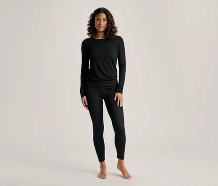 Eine Frau mit dunklem, lockigem Haar steht frontal und trägt ein schwarzes Langarmshirt und schwarze Leggings. Sie ist barfuß.