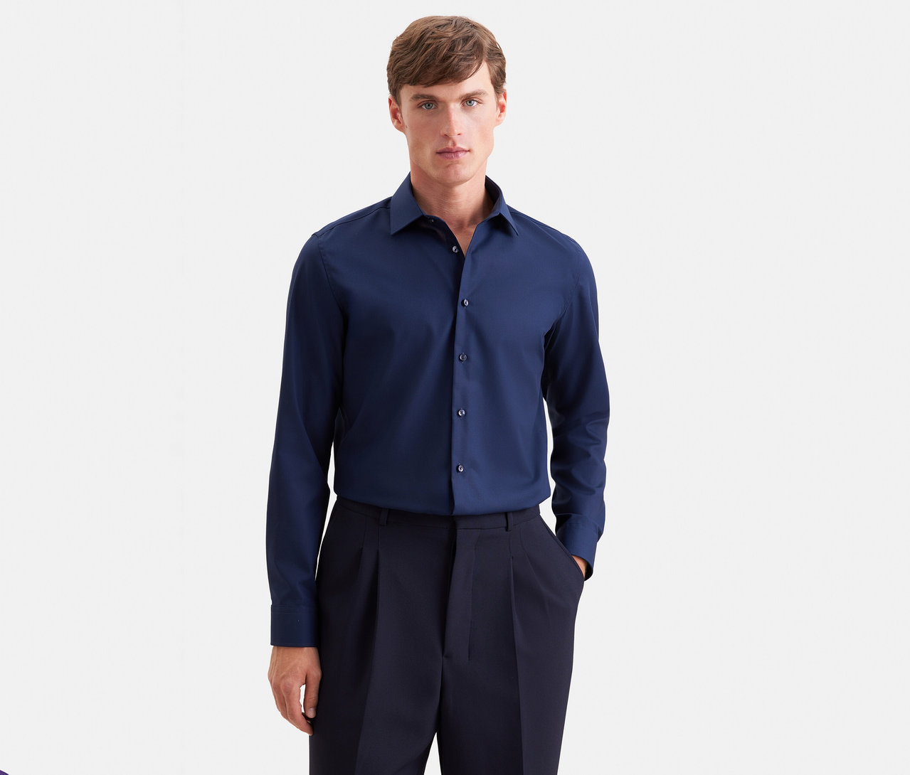 Mann in einem dunkelblauen Langarmhemd und einer dunkelblauen Bundfaltenhose, der nach vorne blickt und eine Hand in der Tasche hat.