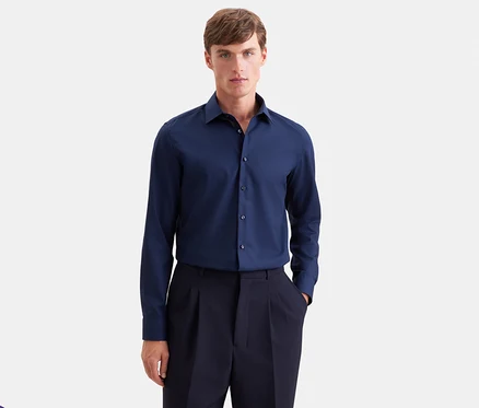 Mann in einem dunkelblauen Langarmhemd und einer dunkelblauen Bundfaltenhose, der nach vorne blickt und eine Hand in der Tasche hat.