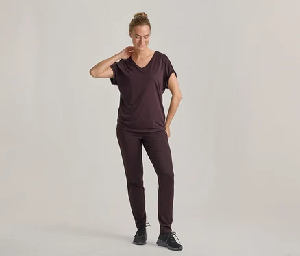 Frau in dunkelbraunem V-Ausschnitt-T-Shirt und passender Hose, mit schwarzen Turnschuhen, steht und blickt nach unten, eine Hand am Nacken.
